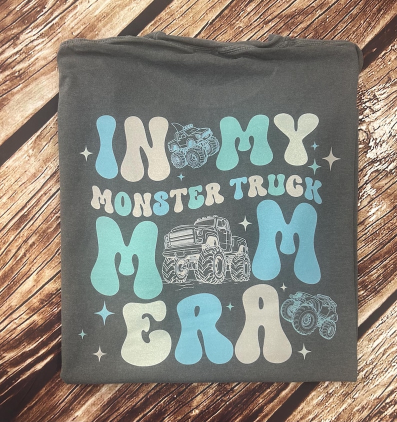 Pu&ograve; includere: T-shirt grigio scuro con la scritta "IN MY MONSTER TRUCK MOM ERA" in bianco e azzurro. Il design include illustrazioni di monster truck e stelle. La maglietta &egrave; piegata su una superficie di legno.