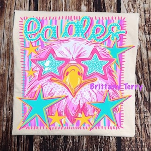 May include: A cream-colored t-shirt featuring a graphic print of an eagle wearing star-shaped teal sunglasses. The design includes the word Eagles in teal, yellow stars, and pink and teal vertical stripes. The text Brittany Terry is printed in pink.