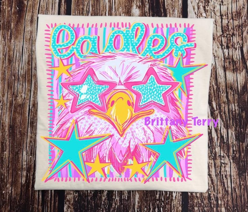 May include: A cream-colored t-shirt featuring a graphic print of an eagle wearing star-shaped teal sunglasses. The design includes the word Eagles in teal, yellow stars, and pink and teal vertical stripes. The text Brittany Terry is printed in pink.