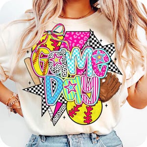 Puede incluir: Camiseta color crema con un gráfico colorido de "Game Day". El diseño incluye una pelota de softbol, un bate de béisbol, un guante y estrellas. El texto está en varios patrones y colores, incluyendo rosa, azul y amarillo.