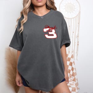 Puede incluir: Una camiseta gris antracita de gran tama&ntilde;o con un n&uacute;mero "3" blanco y una firma roja en la parte delantera.