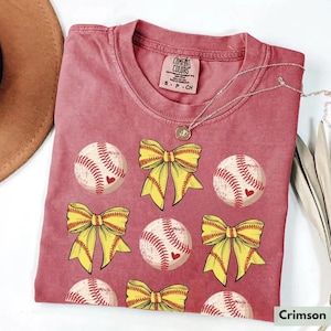 Könnte beinhalten: Karmesinrotes T-Shirt mit einem Muster aus Softbällen mit roten Herzakzenten und gelben Schleifen. Das Shirt hat einen Rundhalsausschnitt und kurze Ärmel. Das Markenetikett ist oben sichtbar. Eine silberne Halskette ist über dem Shirt drapiert.