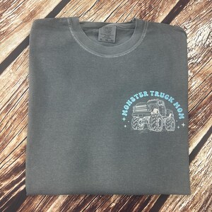 Pu&ograve; includere: Una t-shirt Comfort Colors grigia piegata con scollo tondo. La maglietta presenta una grafica azzurra con la scritta "Monster Truck Mom" e un disegno di un monster truck.