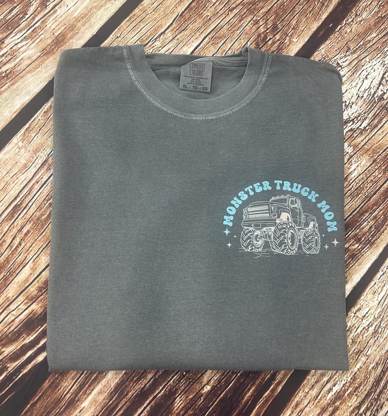 Pu&ograve; includere: Una t-shirt Comfort Colors grigia piegata con scollo tondo. La maglietta presenta una grafica azzurra con la scritta "Monster Truck Mom" e un disegno di un monster truck.