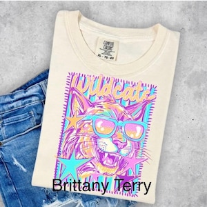 May include: A cream-colored t-shirt featuring a graphic of a wildcat wearing bright blue sunglasses. The design includes the word Wildcats in gold, surrounded by pink and blue stars, all set against a striped background. The shirt has a Comfort Colors label.