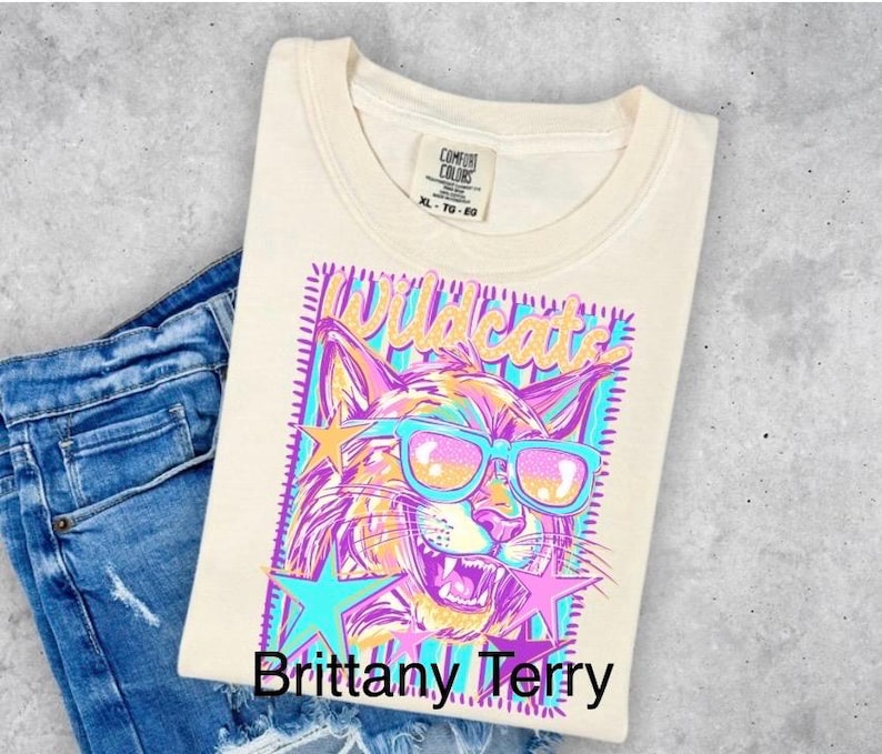 May include: A cream-colored t-shirt featuring a graphic of a wildcat wearing bright blue sunglasses. The design includes the word Wildcats in gold, surrounded by pink and blue stars, all set against a striped background. The shirt has a Comfort Colors label.