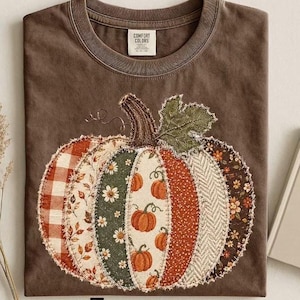 Peut inclure: Un t-shirt marron avec un grand motif central de citrouille composé de divers empiècements en tissu aux motifs variés, notamment à carreaux, floraux et à chevrons. La citrouille possède une tige foncée et une feuille verte.