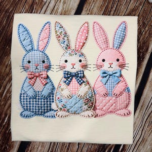 以下が含まれることがあります： クリーム色のシャツに、3匹のアップリケウサギが施されています。各ウサギは、青、ピンク、花柄など、ユニークなデザインです。蝶ネクタイとステッチのディテールがあり、イースターや春に最適です。