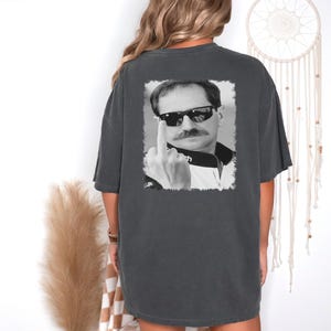 Puede incluir: Una camiseta gris oscuro con una imagen en blanco y negro de un hombre con gafas de sol y haciendo el dedo medio. El hombre lleva una chaqueta de carreras en blanco y negro.