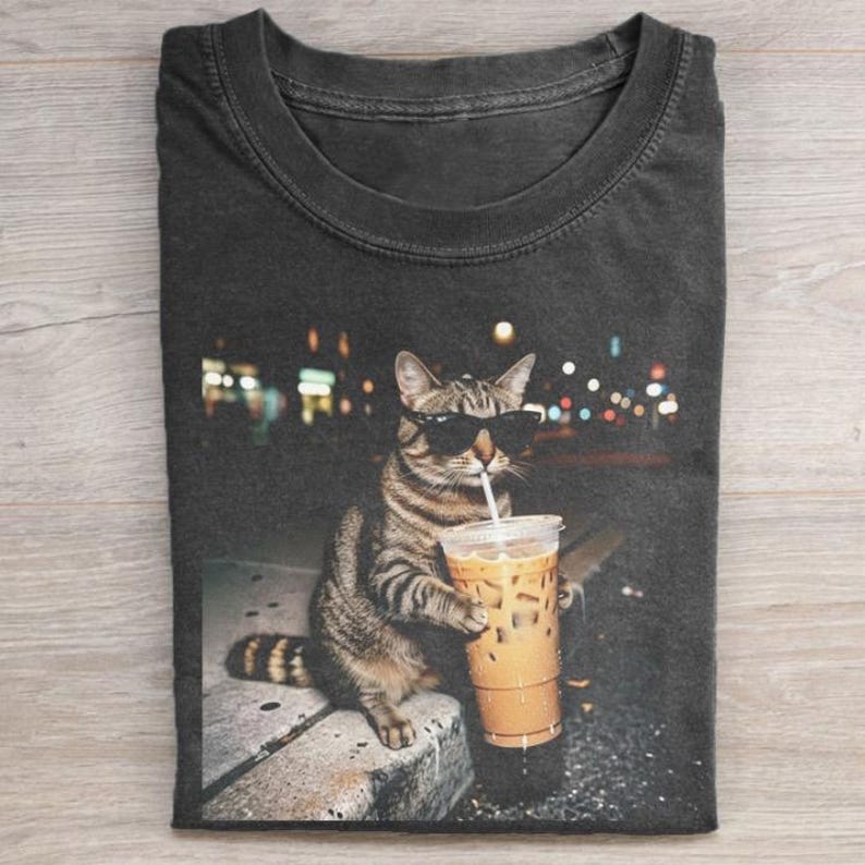 Puede incluir: Camiseta gris oscuro con un gato atigrado con gafas de sol bebiendo caf&eacute; helado. El gato est&aacute; sentado en una repisa con un fondo urbano borroso. La camiseta est&aacute; doblada sobre una superficie de madera.