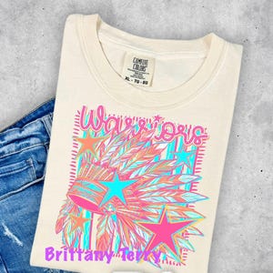 May include: A cream-colored t-shirt featuring a graphic print of a feathered headdress, stars, and the text Warriors and Brittany Terry. The shirt has a Comfort Colors brand label at the neck and is paired with blue denim jeans.