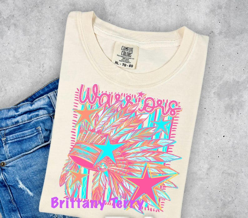 May include: A cream-colored t-shirt featuring a graphic print of a feathered headdress, stars, and the text Warriors and Brittany Terry. The shirt has a Comfort Colors brand label at the neck and is paired with blue denim jeans.