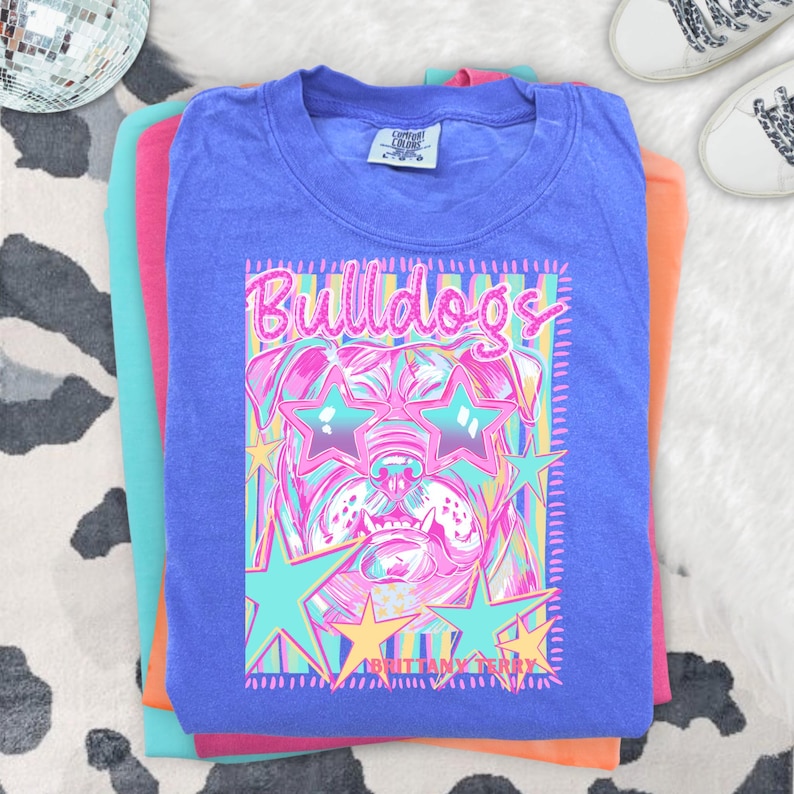 May include: A blue short-sleeve t-shirt featuring a graphic print of a bulldog wearing star-shaped sunglasses. The design includes the text "Bulldogs" and "Brittany Terry" in pink, surrounded by colorful star shapes and vertical stripes on a blue background.