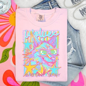 May include: A light pink t-shirt featuring a graphic of a wolf wearing sunglasses. The design includes yellow and blue stars, a striped background, and the text "WOLVES" and "BRITTANY TERRY" in a stylized font. The shirt has a Comfort Colors label.