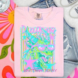 May include: A light pink short-sleeve t-shirt featuring a graphic of a green dragon wearing pink sunglasses. The design includes pink and blue stars, vertical stripes, and the text "Dragons" and "BRITTANY TERRY" in a stylized font.