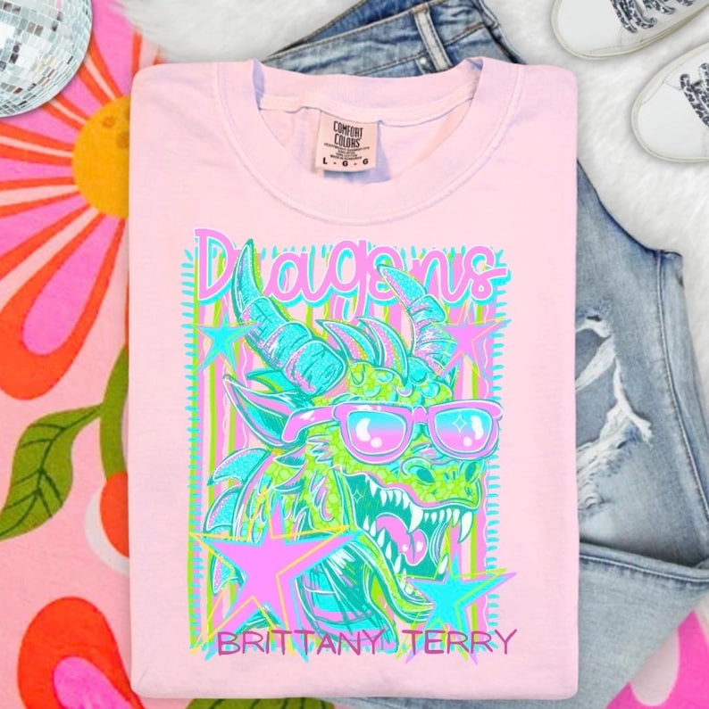 May include: A light pink short-sleeve t-shirt featuring a graphic of a green dragon wearing pink sunglasses. The design includes pink and blue stars, vertical stripes, and the text "Dragons" and "BRITTANY TERRY" in a stylized font.