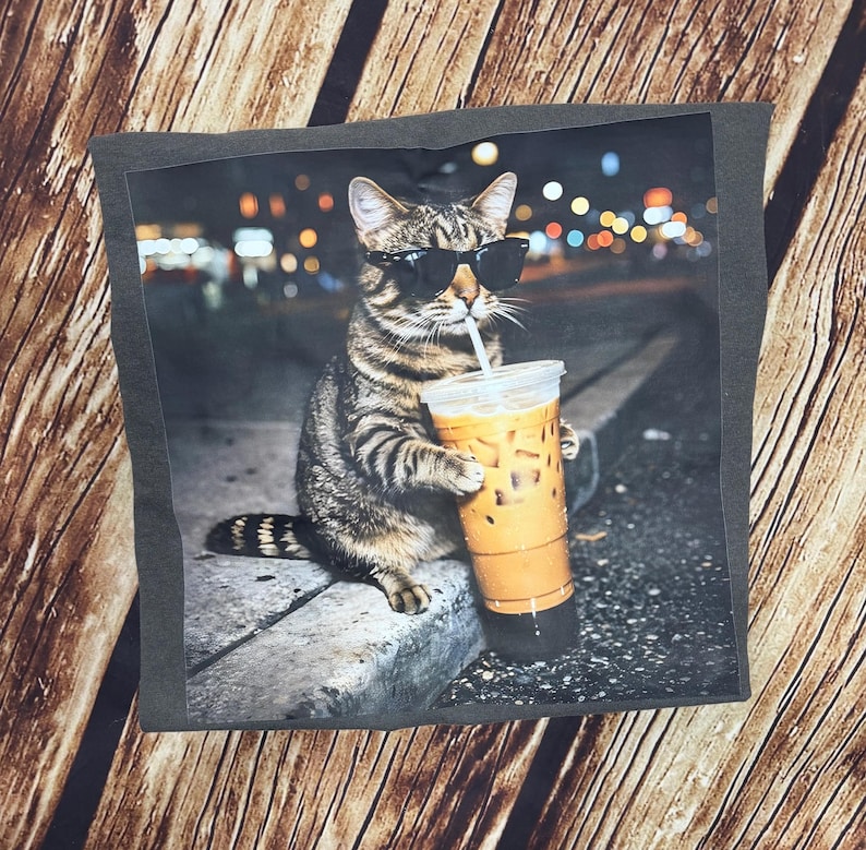 Puede incluir: Una impresi&oacute;n gr&aacute;fica sobre un fondo gris oscuro presenta un gato atigrado con gafas de sol bebiendo caf&eacute; helado. El gato est&aacute; sentado en una acera con un fondo de ciudad. El gato sostiene la taza de caf&eacute; con una pajita.