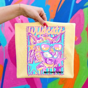 May include: A pale yellow t-shirt featuring a graphic print of a wildcat wearing bright blue sunglasses. The design includes the text "Wildcats" at the top and "BRITTANY TERRY" at the bottom, surrounded by colorful stars and a rectangular border.