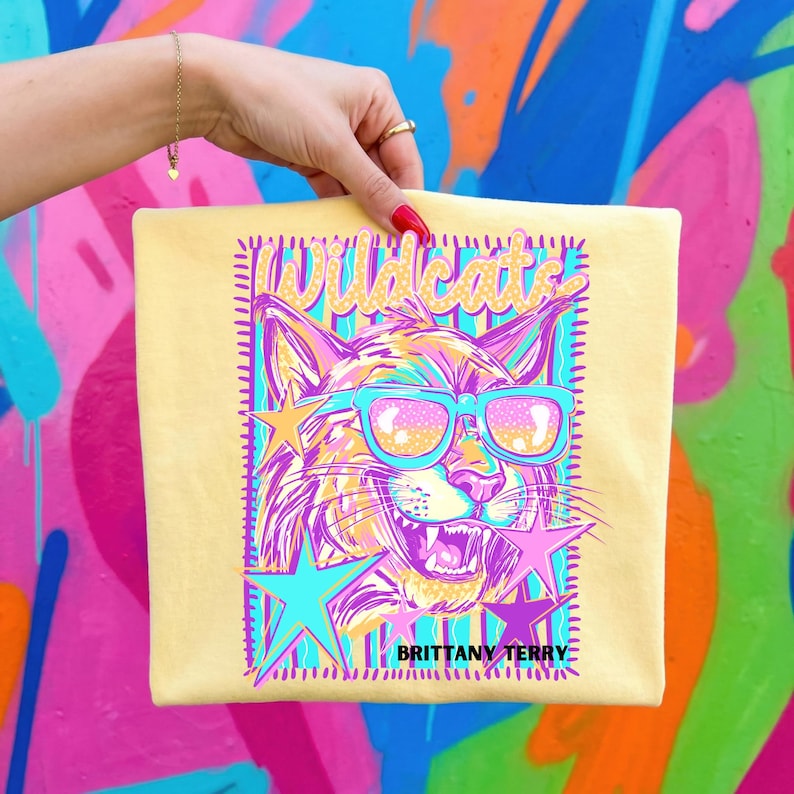 May include: A pale yellow t-shirt featuring a graphic print of a wildcat wearing bright blue sunglasses. The design includes the text "Wildcats" at the top and "BRITTANY TERRY" at the bottom, surrounded by colorful stars and a rectangular border.