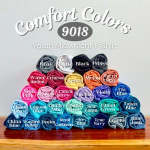 Puede incluir: Una exhibici&oacute;n de camisetas Youth Midweight Comfort Colors enrolladas en varios colores, incluyendo gris, blanco, negro y azul. La imagen presenta el texto "Comfort Colors 9018" y nombres de colores como "Watermelon" y "Orchid".