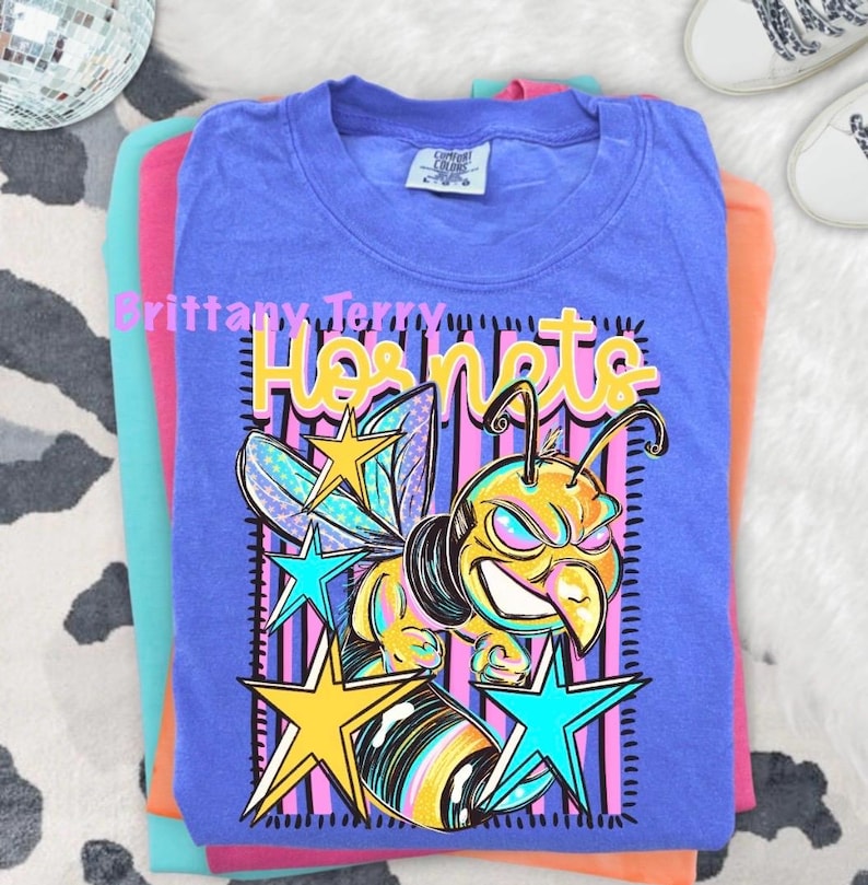 May include: A periwinkle blue t-shirt featuring a graphic of a yellow and black hornet with a fierce expression. The design includes the text Hornets in yellow, surrounded by pink vertical stripes and several colorful stars in yellow and teal.