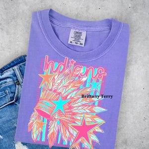 May include: A purple short-sleeve t-shirt featuring a graphic print of a feathered headdress, stars, and the word Indians in a stylized font. The shirt has a Comfort Colors label at the neck and is paired with blue denim jeans.