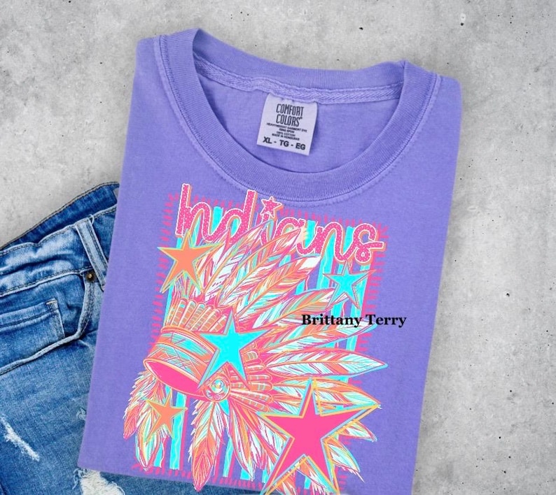 May include: A purple short-sleeve t-shirt featuring a graphic print of a feathered headdress, stars, and the word Indians in a stylized font. The shirt has a Comfort Colors label at the neck and is paired with blue denim jeans.