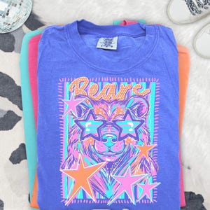 May include: A purple short-sleeve t-shirt featuring a graphic print of a bear wearing star-shaped sunglasses. The design includes the word Bears in orange, surrounded by pink and orange stars against a background of vertical teal and purple stripes.