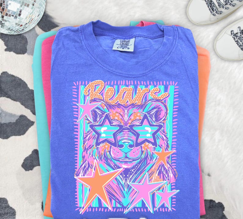 May include: A purple short-sleeve t-shirt featuring a graphic print of a bear wearing star-shaped sunglasses. The design includes the word Bears in orange, surrounded by pink and orange stars against a background of vertical teal and purple stripes.