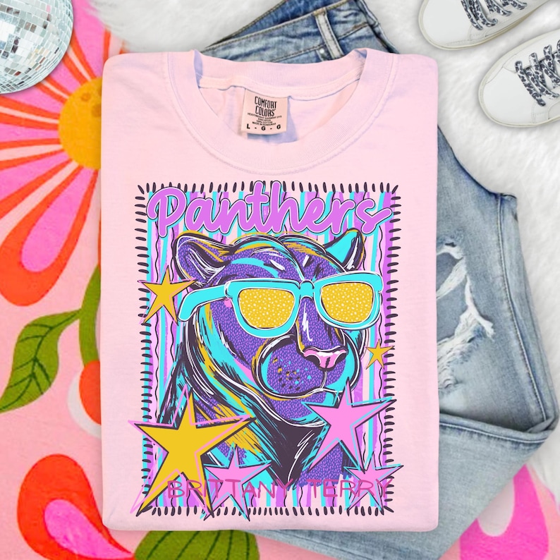 May include: A light pink short-sleeve t-shirt featuring a graphic of a purple panther wearing yellow-lensed teal sunglasses. The design includes the word Panthers in purple, yellow and pink stars, and a striped background with a black dashed border.