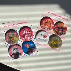 Op de afbeelding: Twee sets knopen met Muppets-personages. Elke verpakking bevat vier knopen met afbeeldingen van Kermit de Kikker, Miss Piggy en andere Muppets. Eén knoop leest "I ❤️ MUPPETS". De verpakking leest "so cute! trinkets".