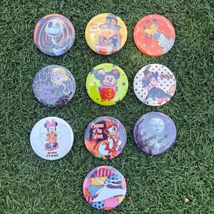 Op de afbeelding: Een verzameling van tien ronde buttons met diverse stripfiguren in Halloween-thema ontwerpen. De buttons tonen kleurrijke illustraties van personages zoals Jack Skellington, Mickey Mouse en Minnie Mouse, inclusief bijbehorende teksten.