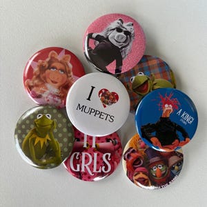 Może przedstawiać: Kolekcja kolorowych, okrągłych przypinek z r&oacute;żnymi postaciami z Muppet&oacute;w. Przypinki zawierają wizerunki Miss Piggy, Kermita Żaby i innych postaci, z napisami takimi jak "I ❤️ MUPPETS" i "A KING!"