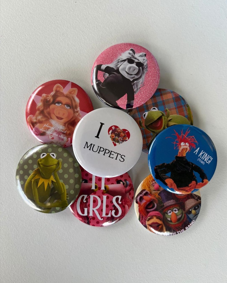 Może przedstawiać: Kolekcja kolorowych, okrągłych przypinek z r&oacute;żnymi postaciami z Muppet&oacute;w. Przypinki zawierają wizerunki Miss Piggy, Kermita Żaby i innych postaci, z napisami takimi jak "I ❤️ MUPPETS" i "A KING!"