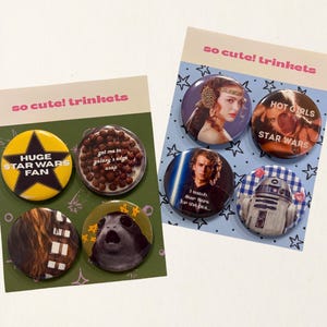 Puede incluir: Dos juegos de chapas circulares en tarjetas de presentación con la etiqueta so cute! trinkets. La tarjeta izquierda incluye chapas con una estrella amarilla, cereales marrones, pelaje de Chewbacca y un Porg. La derecha muestra a Padmé, Anakin y R2-D2.
