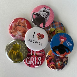 May include: A collection of colorful round buttons featuring various Muppets characters. The buttons showcase images of Miss Piggy, Kermit the Frog, and other characters, with text like "I ❤️ Muppets" and "A King!"