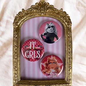 Może przedstawiać: Złota, ozdobna rama zawiera trzy okrągłe guziki z Miss Piggy. Guziki są r&oacute;żowe i czerwone z wizerunkami Miss Piggy oraz napisami "IT GRLS" i "please do not disturb the star". Tło jest w r&oacute;żowo-białe paski.