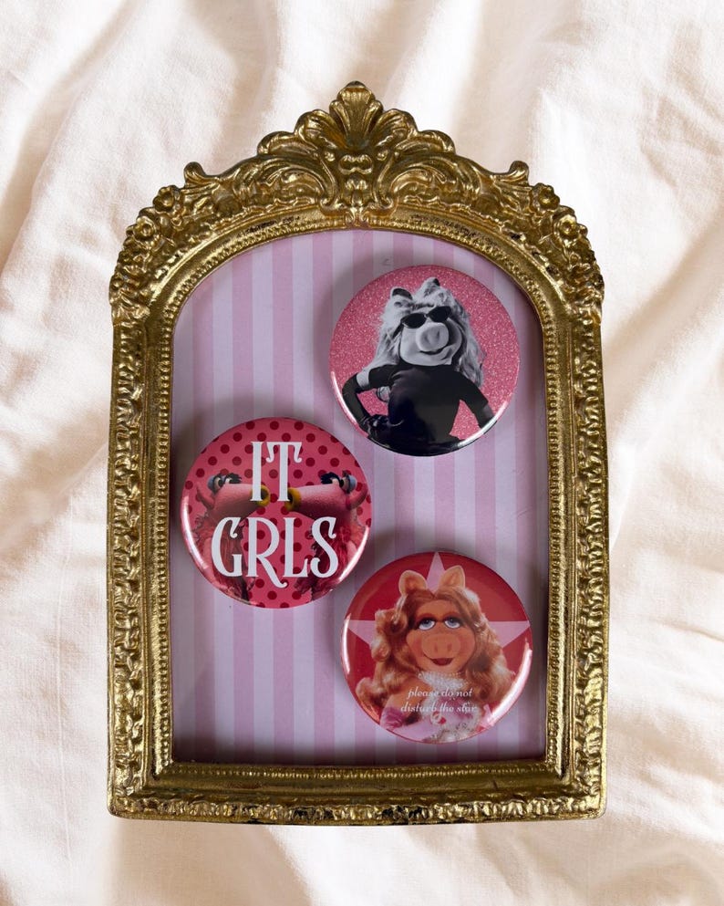 Może przedstawiać: Złota, ozdobna rama zawiera trzy okrągłe guziki z Miss Piggy. Guziki są r&oacute;żowe i czerwone z wizerunkami Miss Piggy oraz napisami "IT GRLS" i "please do not disturb the star". Tło jest w r&oacute;żowo-białe paski.