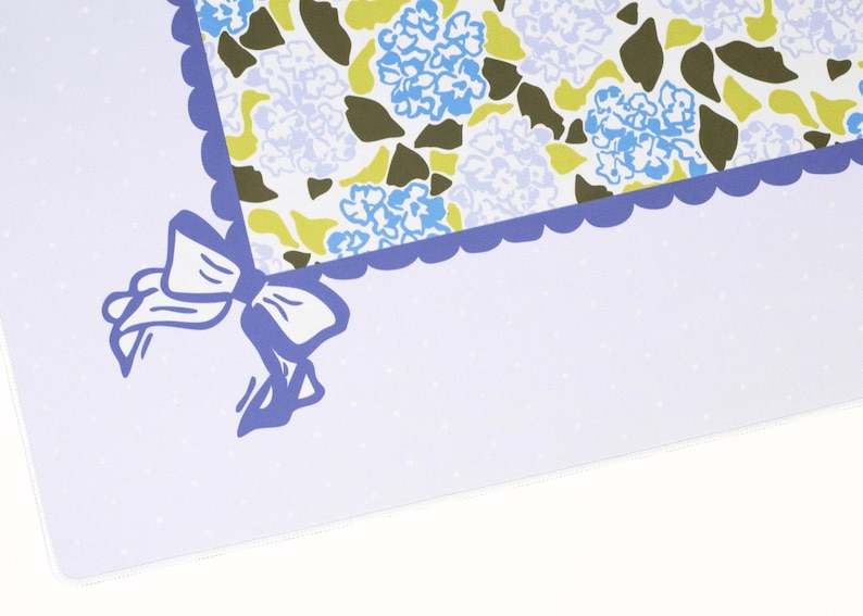 May include: A decorative paper with a floral pattern featuring blue hydrangeas, green leaves, and a scalloped border. A blue bow is in the corner. The background is white with small, light blue polka dots.