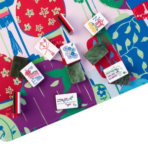 Może przedstawiać: Kolorowy zestaw do gry w Mahjonga z kafelkami rozrzuconymi na wzorzystej macie. Mata ma żywy wz&oacute;r z czerwonymi, fioletowymi i niebieskimi elementami. Kafelki są białe z r&oacute;żnymi symbolami i cyframi, a także obecne są zielone uchwyty na kafelki.