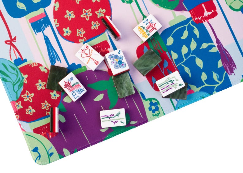 Może przedstawiać: Kolorowy zestaw do gry w Mahjonga z kafelkami rozrzuconymi na wzorzystej macie. Mata ma żywy wz&oacute;r z czerwonymi, fioletowymi i niebieskimi elementami. Kafelki są białe z r&oacute;żnymi symbolami i cyframi, a także obecne są zielone uchwyty na kafelki.