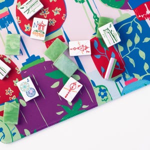 Może przedstawiać: Kolorowy zestaw do gry w Mahjonga z kafelkami i zielonymi, czerwonymi i fioletowymi akcentami. Gra jest ustawiona na wzorzystej macie z kwiatowymi i geometrycznymi wzorami. Kafelki są białe z r&oacute;żnymi symbolami.