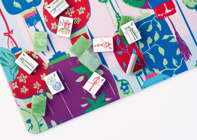 Może przedstawiać: Kolorowy zestaw do gry w Mahjonga z kafelkami i zielonymi, czerwonymi i fioletowymi akcentami. Gra jest ustawiona na wzorzystej macie z kwiatowymi i geometrycznymi wzorami. Kafelki są białe z r&oacute;żnymi symbolami.