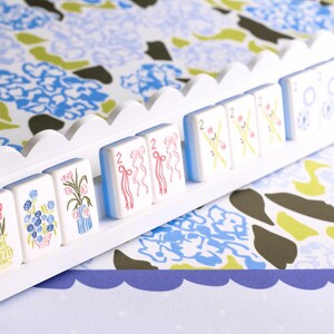 May include: A white wooden rack holding decorative tiles with floral and ribbon designs. The tiles are white with blue, pink, and green illustrations. The background features a floral pattern in blue, green, and white.