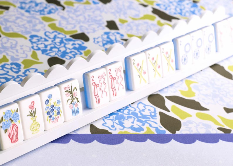 May include: A white wooden rack holding decorative tiles with floral and ribbon designs. The tiles are white with blue, pink, and green illustrations. The background features a floral pattern in blue, green, and white.