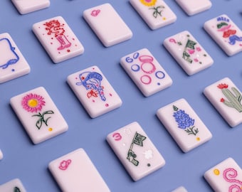 May include: A collection of white rectangular mahjong tiles arranged in a grid pattern on a light purple surface. Each tile features a unique, colorful hand-painted illustration, including flowers, a cowboy hat, a gun with a flag, and geometric shapes.
