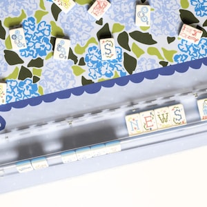 May include: A close-up of a light blue and white floral patterned surface with scattered game tiles. A clear plastic holder contains tiles spelling "NEWS". The tiles have various symbols and numbers.