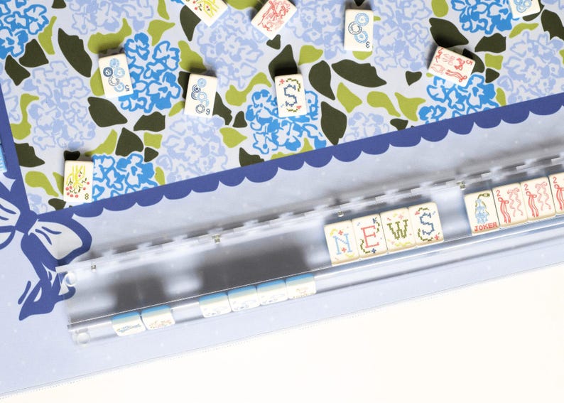May include: A close-up of a light blue and white floral patterned surface with scattered game tiles. A clear plastic holder contains tiles spelling "NEWS". The tiles have various symbols and numbers.