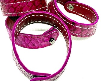 Può includere: Tre braccialetti in pelle magenta con cuciture bianche e chiusure a scatto argentate. I braccialetti hanno un motivo strutturato simile alla pelle di serpente. L'interno dei braccialetti è di un colore bordeaux più scuro.