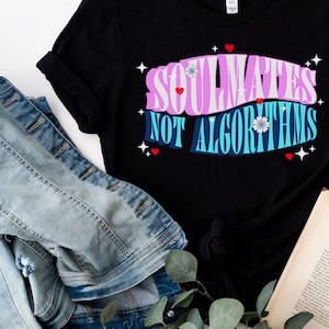 May include: Black t-shirt with the text "Soulmates Not Algorithms" in pink, blue, and white lettering with stars and hearts. The shirt is laid flat on a white surface with a denim jacket, a book, a hat, and greenery.